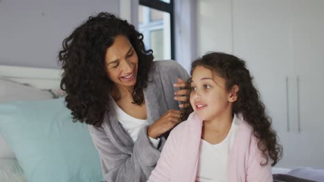 Happy-mixed-race-mother-and-daughter-sitting-in-bedroom-and-playing-with-hairs