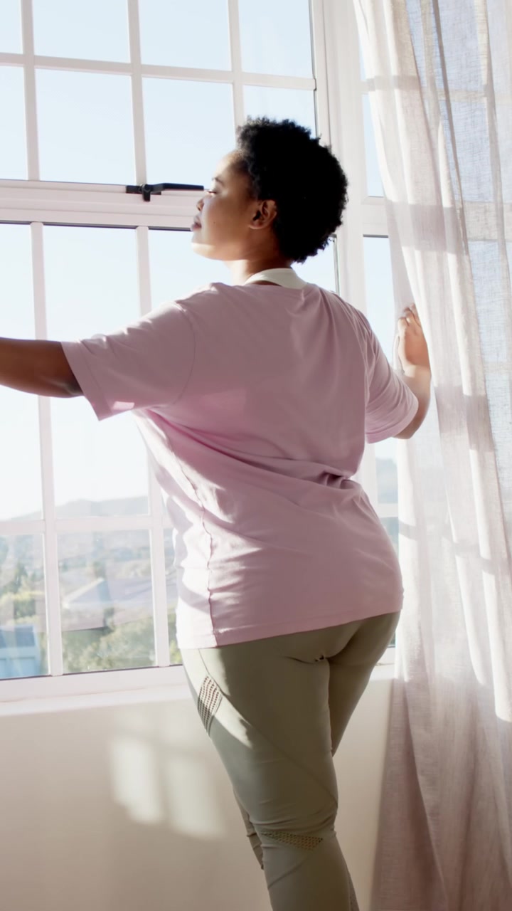 Video verticale: Donna afroamericana attratta dal sole, che tira le tende velate alla finestra, osservando.