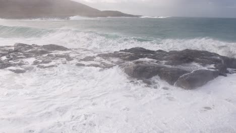 panning, slow motion shot of a large, white wave breaking over the rocks during a storm in the bay by tangasdale beach, near castlebay on the isle of barra