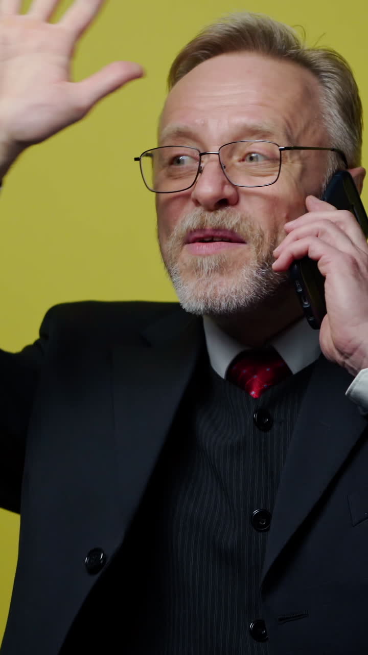 Happy senior businessman talking the phone. Portrait of a mature man in black costume speaking on the mobile phone and gesticulating a lot isolated on yellow. Vertical video
