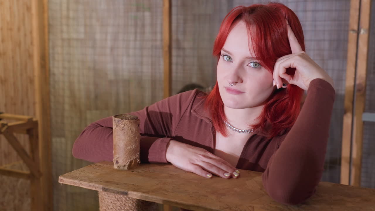 redhaired woman smiling at camera while leaning on cat tree, warm bamboo backdrop and soft studio light playful mood with easy laughter, necklace detail, friendly glance and relaxed hands on wooden