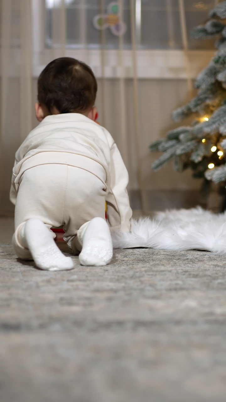 Little child in white clothes crawls by the room followed by camera. Baby stops at the toy pyramid near the Christmas tree. Vertical video