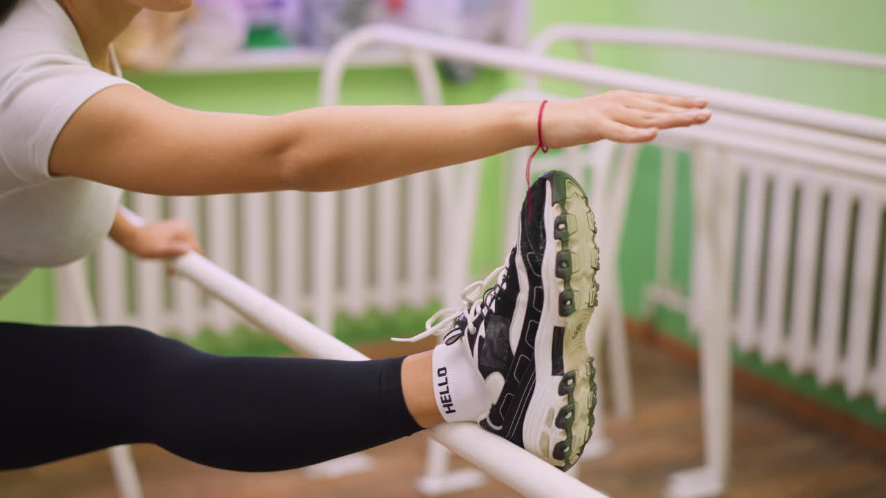 Lady in sportswear stretching leg on white bars indoors, black sneakers with white socks visible, arm extended for balance, exercise showing focus and flexibility in a fitness training environment
