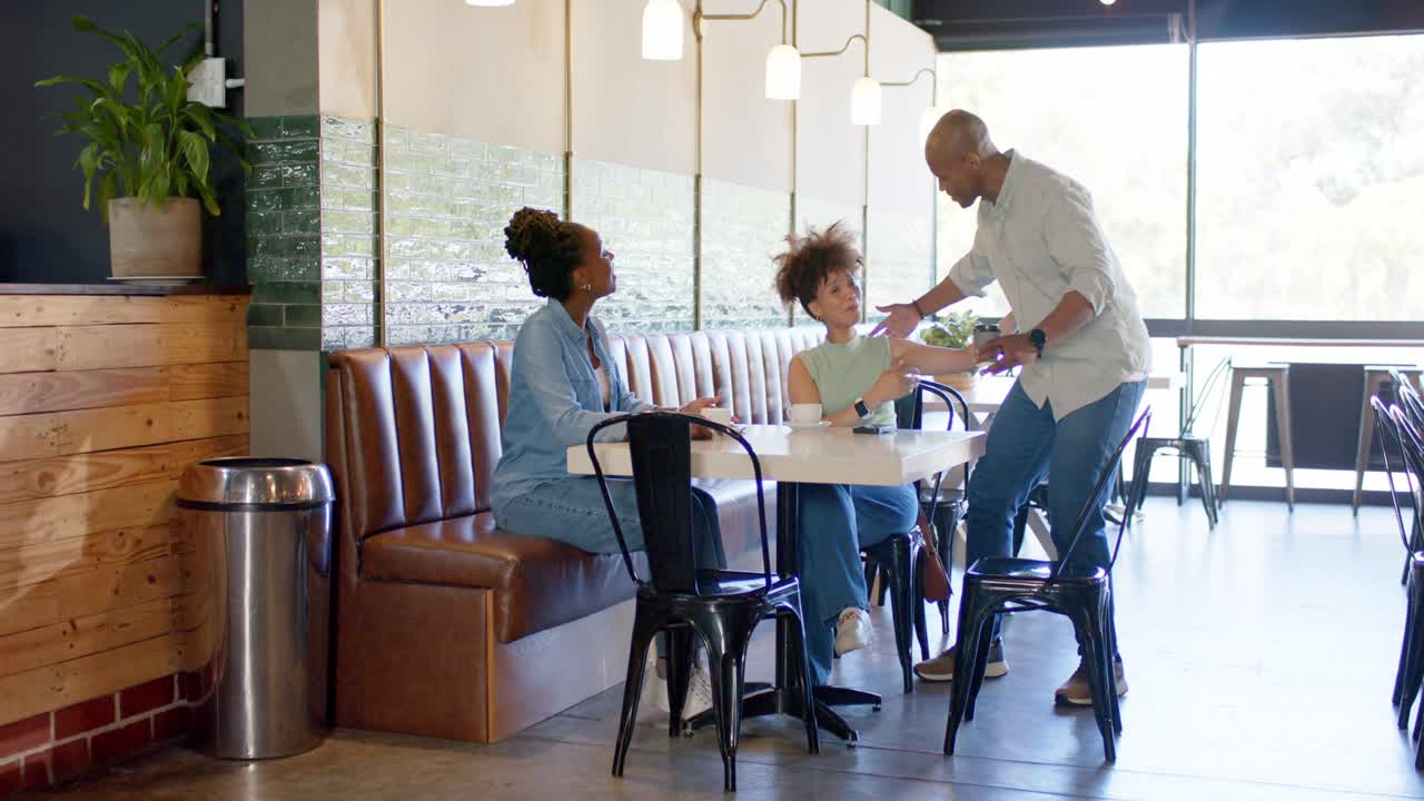 Man is entering and offering takeaway cup at booth table near bright window, greeting women