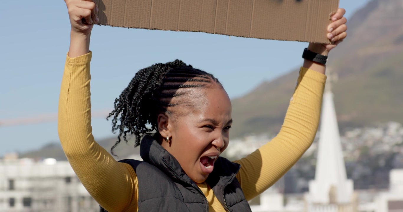 Femme afro-américaine vêtue d'un haut jaune brandissant une pancarte en carton 'FEMMES' sur une colline, criant pour se faire entendre.