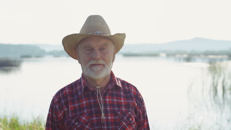 Portrait-of-a-senior-haired-man-in-hat-smiling-at-the-camera-on-the-picturesque-lake-background