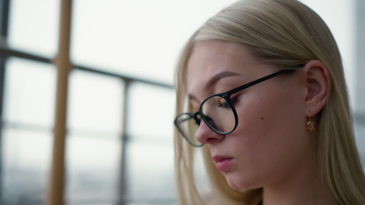 primer plano de una mujer pensativa con gafas, sosteniendo una pluma en la boca mientras mira hacia afuera a través de un panel de vidrio, su expresión pensativa destaca un momento de reflexión en un entorno acogedor