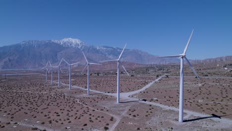 Statische-Drohnenaufnahme-Erfasst-Windmühlen-In-Der-Wüste-Mit-Schneebedeckten-Bergen-Und-Weiteren-Windmühlen-Im-Hintergrund