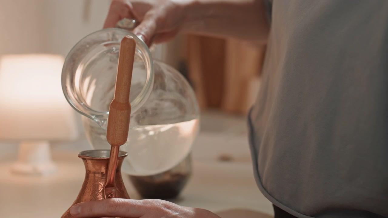Mani in primo piano che versano acqua in un bollitore di vetro con base in rame e fondi di caffè sfocati, bagliore di una lampada sullo sfondo, versamento lento e preciso, vapore caldo e dettagli tattili che catturano un tranquillo rituale sensoriale