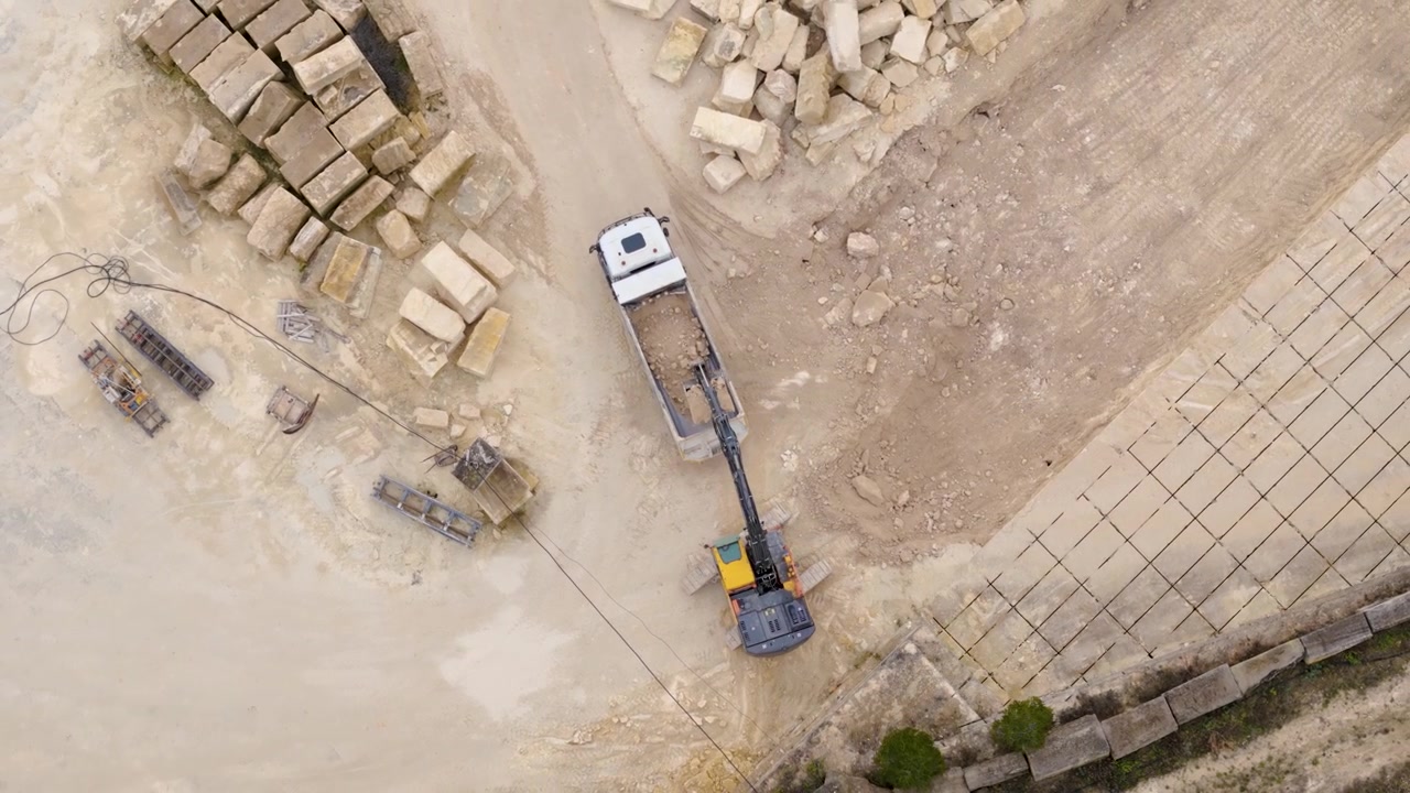 En droneoptagelse ovenfra af en gravemaskine, der fylder en dumper med kalksten eller lyst beige sand