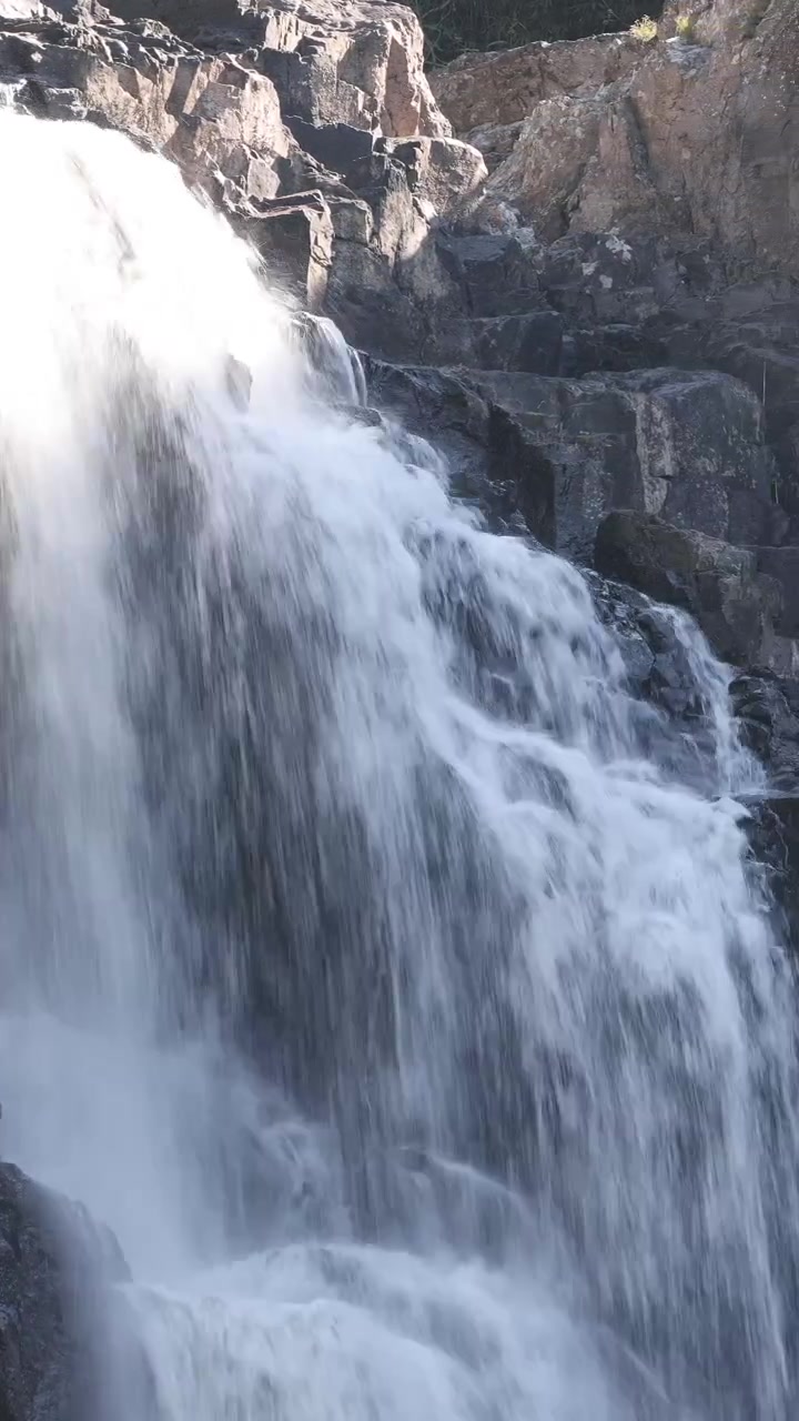 Cascata impetuosa scorre su rocce scoscese, luce diurna intensa, telecamera stabile, ambiente esterno naturale