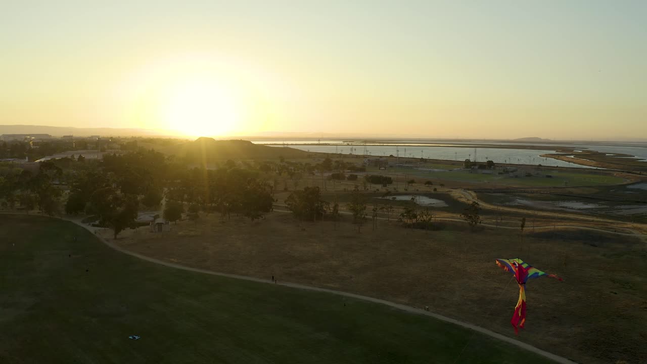 Aerial drone shot of high flying kite, drone slowly moves in to bright colorful kite as it weave and zips around, floating with sunset and wetlands on horizon in background