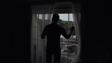 Handsome-groom-in-gray-jacket-goes-to-balcony-and-opens-curtains-in-hotel-room-or-home-apartment