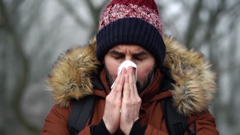 bearded caucasian male places white paper tissue over his face and blows his nose in forest during cold winter day, slow motion