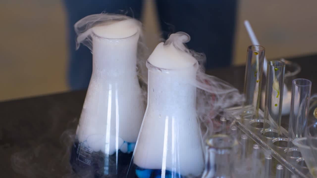 Expérience de laboratoire spectaculaire avec plusieurs fioles Erlenmeyer projetant violemment une épaisse fumée blanche de glace carbonique sur des liquides bleus et jaunes dans un portoir à éprouvettes, entourée de verrerie scientifique.