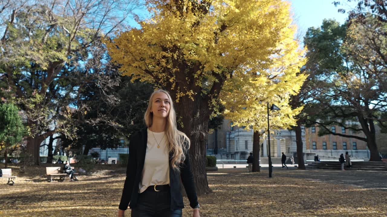 A serene autumn scene in Tokyo, where a woman strolls through a park filled with vibrant yellow foliage.