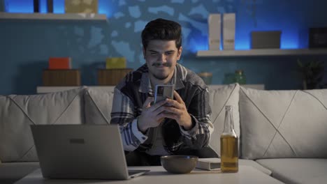 hombre usando el teléfono y bebiendo alcohol en casa por la noche.