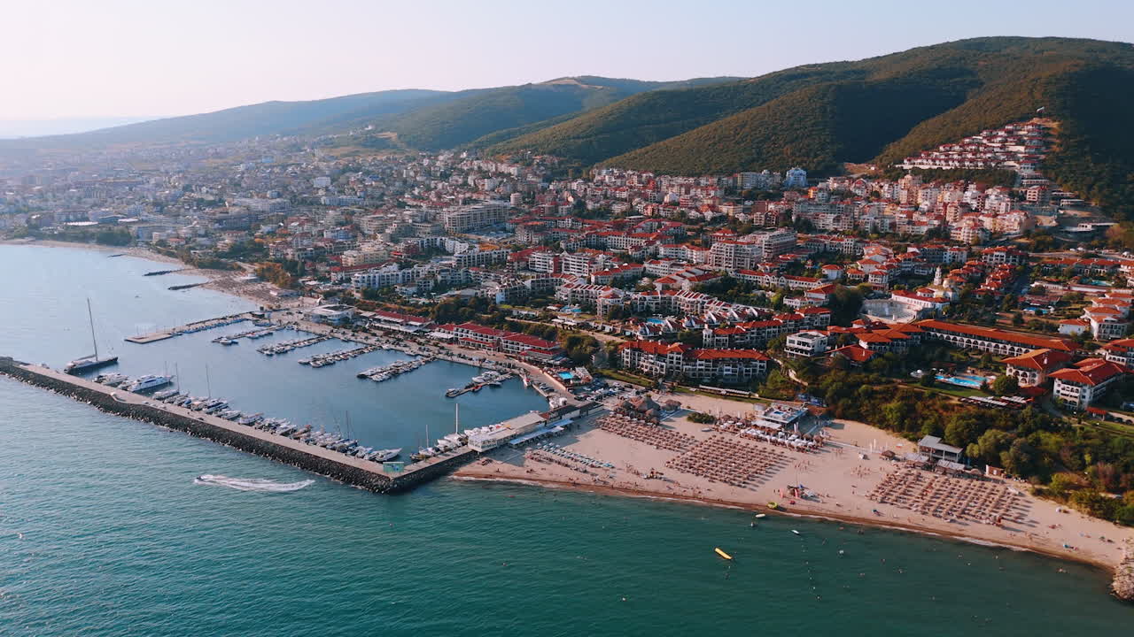Muhteşem marina ve plaj manzarası. Tepeler ve renkli binalarla çevrili, berrak gökyüzünün altında canlı bir marina ve kumlu plajın havadan görünümü