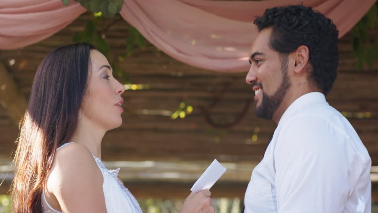 Diverse couple at pink drape arbor bride dress reading vows groom smiling hands camera pulling back