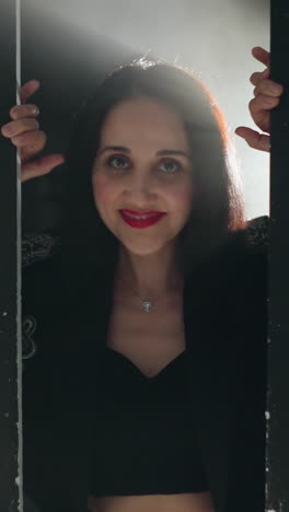mujer serena en traje negro enmarcada por una puerta, bañada en luz suave con un zoom lento de la cámara mientras sonríe suavemente