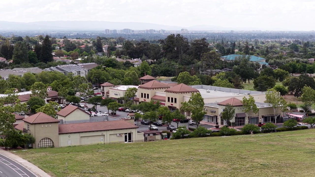 Vista di tracciamento con drone che sorvola l'Evergreen Valley College e un centro commerciale a San Jose, California, con il lussureggiante paesaggio della Silicon Valley che si estende verso lo skyline della città in lontananza.
