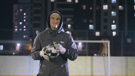 atleta con capucha y guantes sosteniendo una pelota de fútbol con una sonrisa sutil en el campo deportivo urbano por la noche, el fondo muestra edificios altos iluminados, postes de portería y efectos de luz bokeh suaves