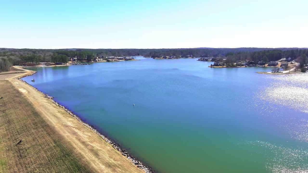 Lake Treasure with sun reflection in Georgia during sunny day. Forest landscape with small village named Fairfield Plantaiton. Dam with rocky shore. Descend wide shot.