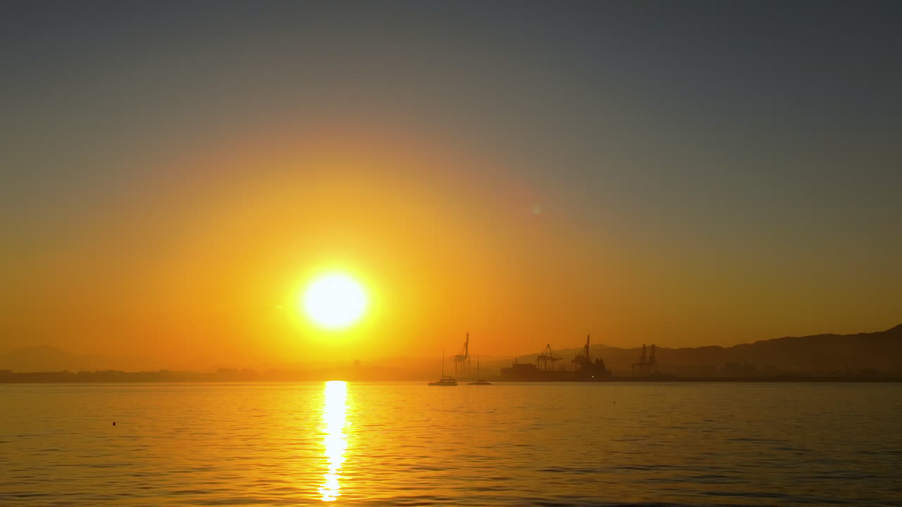 Artistic look of sunset over the ocean, with an orange sky and silhouettes of distant structures on the horizon reflecting over the calm sea surface
