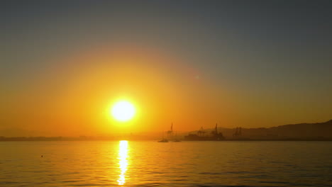 artistic look of sunset over the ocean, with an orange sky and silhouettes of distant structures on the horizon reflecting over the calm sea surface