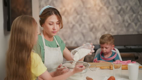fair skinned girl holds sieve as her mum pours flour into bowl during baking session while boy in background wears pink toy rings on neck and wrist playing with cookie cutter at kitchen table