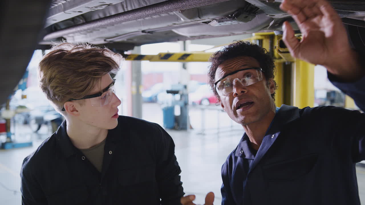 Male Tutor With Student Looking Underneath Car On Hydraulic Ramp On Auto Mechanic Course