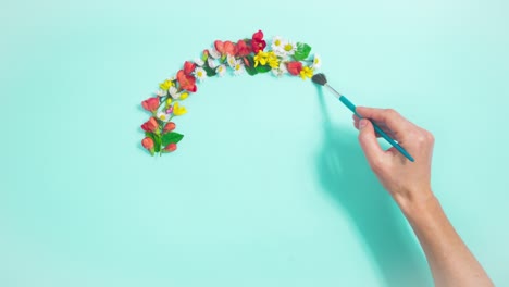 woman's hand draws circle with brush from natural flowers, buds and young leaves on light blue background with copy space in center.