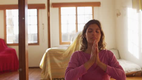 Relaxed-mixed-race-woman-practicing-yoga,-sitting-meditating-and-smiling-in-sunny-cottage-bedroom