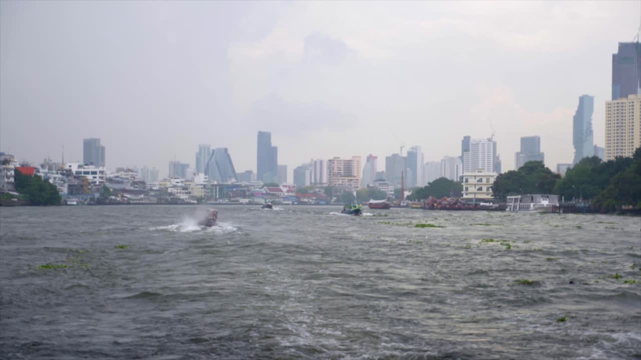 Slow motion shot of the Chao Phraya River in Bangkok taken from a boat. Other boats are visible on the water and the skyscrapers of the city are visible in the background