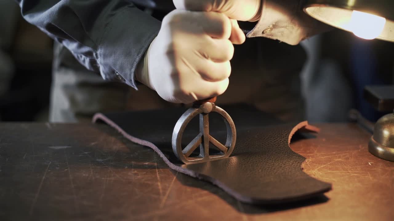Grasping handle, gloved hands pressing peace stamp into leather for branding at bench, lamp glow