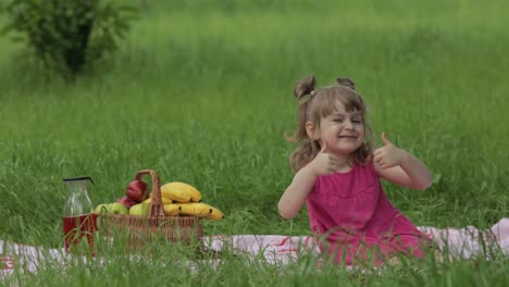 Weekend-at-picnic.-Lovely-caucasian-child-girl-on-green-grass-meadow-sit-on-blanket-show-thumbs-up