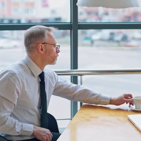 mature man in white shirt sitting at the table near the window and working on a laptop. businessman with coffee enjoys city background through the window.