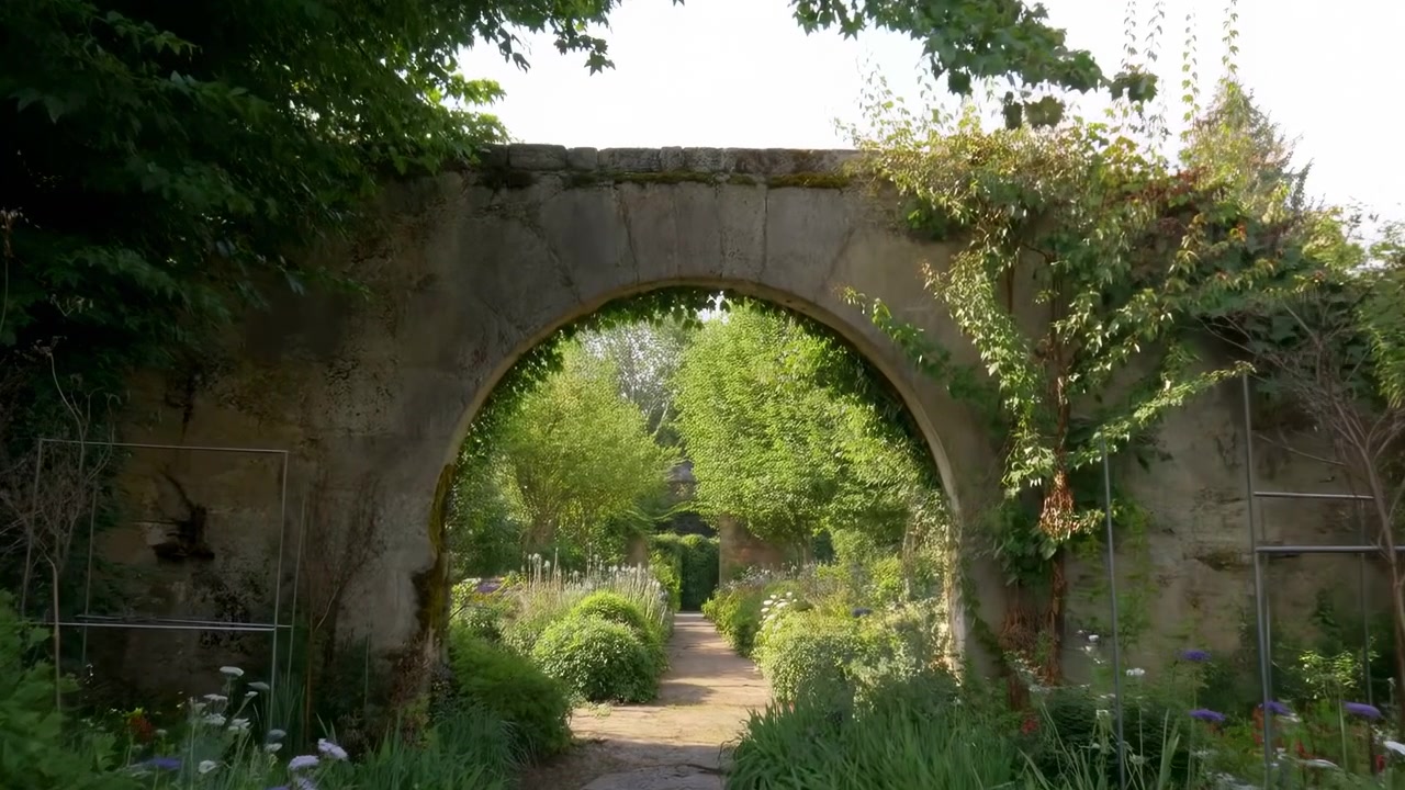 Ancient stone arch remains centered while leafy plants surround pathway in soft daylight, showing layered greenery, curved masonry texture, and peaceful composition from front viewpoint.