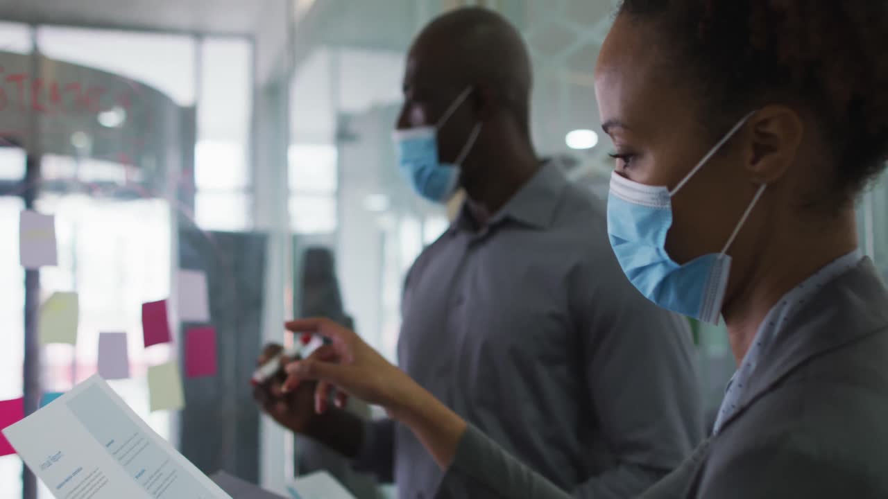 Diverse male and female business colleagues in face masks discussing by transparent board in office