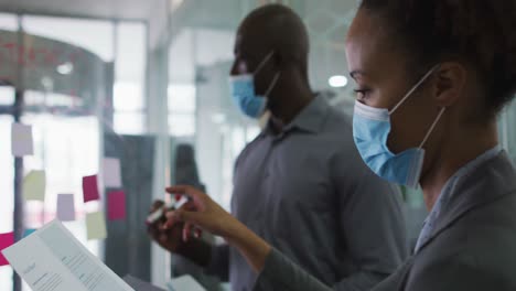 diverse male and female business colleagues in face masks discussing by transparent board in office