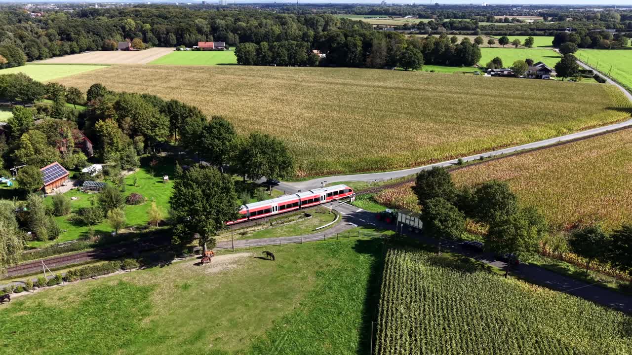 Aerial tracking shot of red train on railway in countryside. Sunny summer day with forest trees and cultivated farmland fields. Orbit wide shot