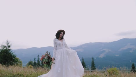 Happy bride on mountains background. Luxury woman in beautiful wedding dress holds flowers and posing on camera among natural landscape.