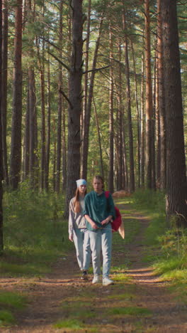 dos amigos caminando por un bosque pacífico rodeado de árboles altos, uno lleva una bufanda azul y sigue detrás, mientras que el otro en verde conduce con una mochila roja drapeada con un pañuelo