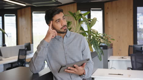 Using tablet, Indian man standing in modern office, focusing on work tasks