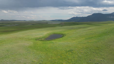 Valle-De-Pradera-De-Hierba-Verde-Con-Un-Pequeño-Lago-Con-Luz-Solar-Atravesando-Un-Día-Semi-Nublado-Y-Montañas-Oscuras-Al-Fondo-[imágenes-De-Drones-De-4k