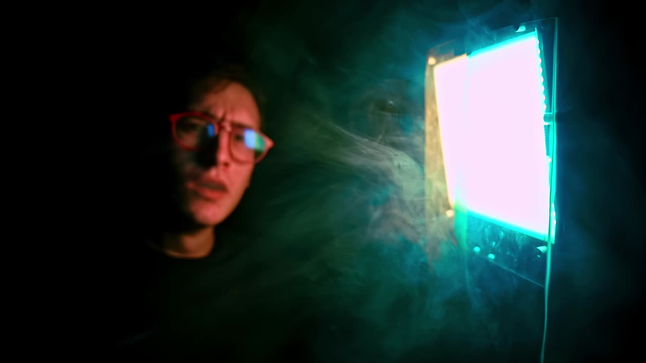 Young serious man in red glasses vaping in front of a green yellow light source in a dark room. Side view. Close up. Slow motion