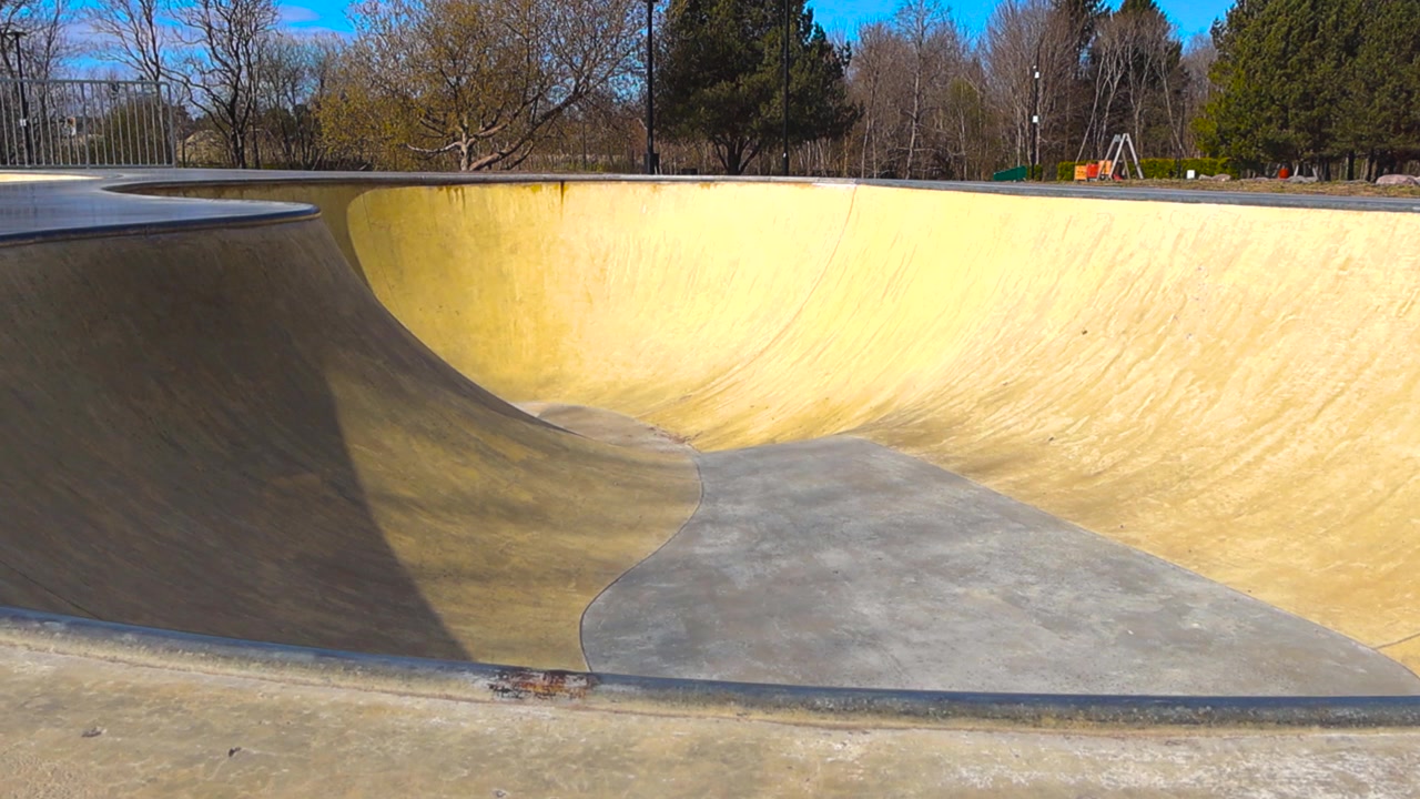 Movimento fluido della telecamera che rivela una vasca da skatepark in cemento giallo e grigio, completamente vuota in una giornata luminosa e soleggiata con alberi sullo sfondo, perfetta per sport estremi e attività ricreative urbane