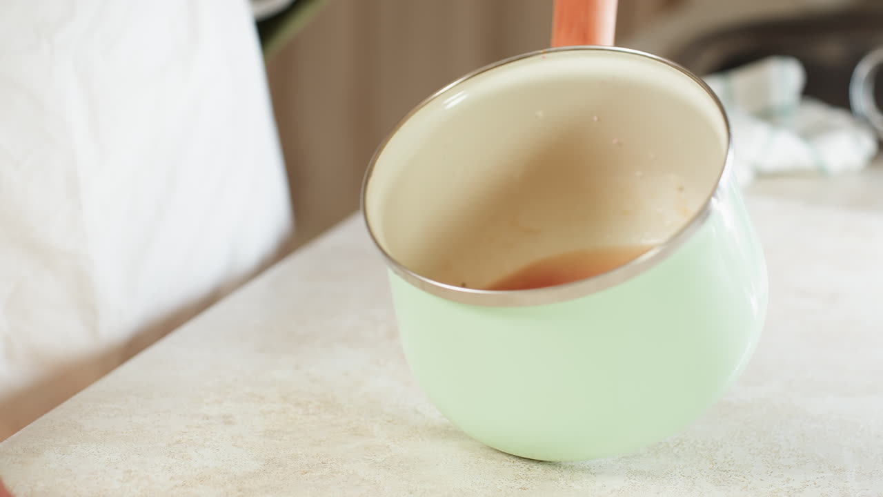 Close up of person holding mint green cooking pot by wooden handle and stirring contents inside over kitchen counter, showing gentle hand movement in bright indoor kitchen