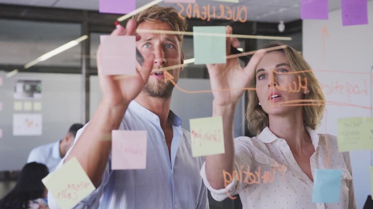 Professionisti, un uomo e una donna, che discutono di promemoria su una lavagna in vetro in un ufficio moderno.