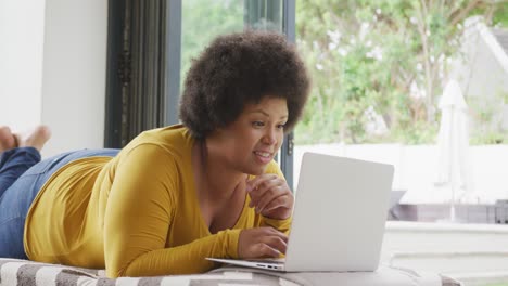 video de una feliz mujer afroamericana de tamaño grande acostada en el suelo y usando una computadora portátil